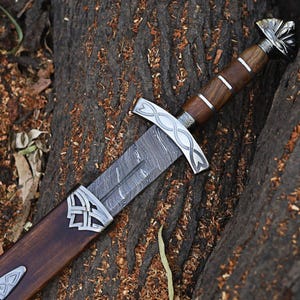 May include: A sword with a patterned Damascus steel blade, a dark brown wooden handle with silver-colored metal rings, and a silver-colored crossguard featuring an infinity knot design. The sword is partially inserted into a matching wooden scabbard.