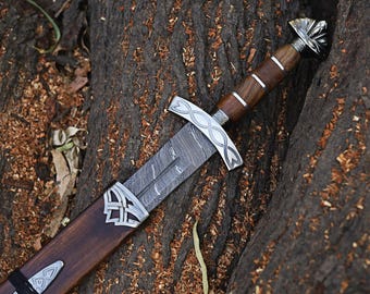 May include: A detailed image of a sword with a patterned blade and a wooden handle. The sword is in a wooden sheath. The handle has silver accents. The sword is resting on a tree trunk.