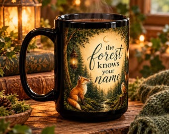 May include: A black ceramic mug featuring a printed forest scene with a fox, an owl, a hanging lantern, and the text 'the forest knows your name' in black script. The design includes a crescent moon above a dense woodland landscape.