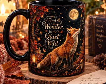 May include: A black ceramic mug featuring a graphic of a fox standing in a forest at night under a full moon. The text reads Find Wonder in the Quiet Wild. The design includes autumn leaves, a lantern, and mushrooms in shades of orange and brown.