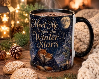 May include: A ceramic mug with a black handle and rim, featuring a dark blue night scene with two foxes wrapped in a blanket, a full moon, and snowy trees. The text reads Meet Me Under the Winter Stars in gold script lettering.