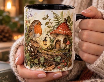 May include: A white ceramic mug with a black interior and handle, featuring a detailed illustration of a robin perched on a tree stump next to a small mushroom-shaped house, a hanging lantern, and various forest plants and flowers.
