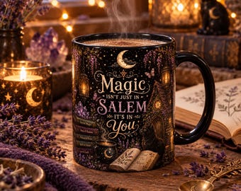 May include: A black ceramic mug featuring a crescent moon, a small house, a black cat, and an open book. The text reads: Magic ISN'T JUST IN SALEM IT'S IN You. The mug is filled with a steaming beverage and displays illustrations of books, lanterns, and purple flowers.