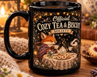 May include: A black ceramic mug featuring a colorful illustration of a fox and a badger having tea. The text reads Official Cozy Tea & Biscuit Society. The scene includes a table with a teapot, cups, and biscuits, surrounded by flowers and fairy lights.