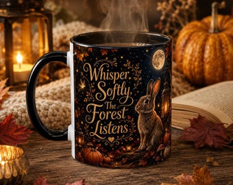 May include: A black ceramic mug featuring a graphic of a brown rabbit, a full moon, and pumpkins. The text reads, Whisper Softly, The Forest Listens. Steam rises from the top of the mug, which is decorated with autumn leaves and golden vines.