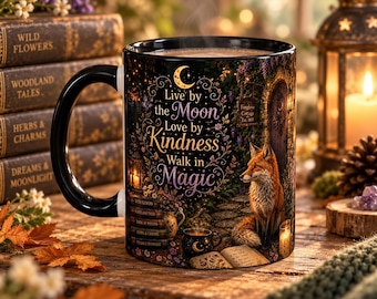 May include: A black ceramic mug featuring a printed illustration of a fox sitting on a stone path near a cottage door. The text reads: Live by the Moon, Love by Kindness, Walk in Magic. The design includes a crescent moon, flowers, and books.