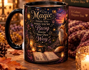 May include: A black ceramic mug featuring a colorful illustration of a fox in a forest setting with lanterns, an open book, and the text: Magic FINDS YOU WHEN YOU'RE Busy MAKING YOUR OWN Way. The book pages read: Believe in the magic within and Tiny steps still lead to big things.