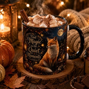 May include: A black ceramic mug filled with hot cocoa, marshmallows, and a cinnamon stick. The mug features a printed illustration of a fox in an autumn forest with the text Find Magic in the Quiet Moments, set against a dark starry night sky.