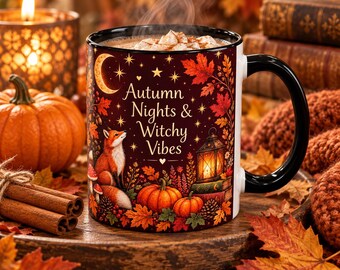 May include: A ceramic mug with a black handle and rim, featuring a dark red autumn-themed illustration of a fox, pumpkins, a lantern, and stars. The text reads Autumn Nights & Witchy Vibes. The mug is filled with a warm beverage topped with foam.