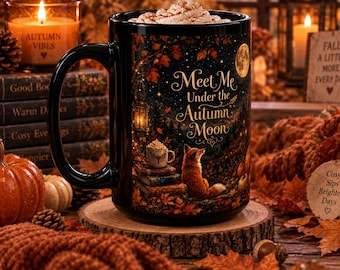 May include: A black ceramic mug featuring a printed illustration of a fox sitting near a stack of books and a lantern under a full moon. The text "Meet Me Under the Autumn Moon" is printed in gold script. The mug is filled with whipped cream.