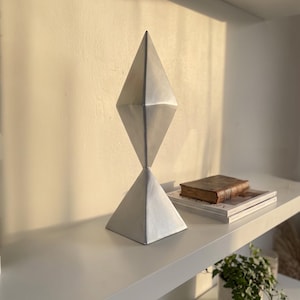 Pode incluir: Uma escultura de cor prateada com um design geométrico, apresentando uma secção superior em forma de diamante sobre uma base de pirâmide quadrada. O objeto tem um acabamento mate liso e mantém-se na vertical com arestas bem definidas.