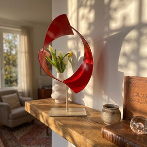 Pode incluir: Uma escultura de metal vermelho com um design curvo e circular montada numa base quadrada, posicionada atrás de um vaso de cerâmica branca com tulipas brancas e amarelas. A escultura apresenta um acabamento vermelho sólido e um suporte fino.