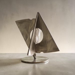 May include: A brushed metal sculpture featuring two intersecting triangular planes mounted on a circular base. The structure includes a central circular cutout and small notches along the edges, creating a geometric form with a matte finish.