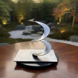 Pode incluir: Uma escultura de metal moderna e abstrata com um design em forma de fita rodopiante. A escultura prateada assenta numa base quadrada de metal escovado, montada numa base circular preta. A escultura é exibida numa mesa de madeira escura.