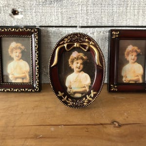 May include: Three ornate picture frames with gold accents. The frames are all different shapes and sizes. Each frame contains a black and white photograph of a young girl.
