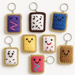 May include: Handmade crochet keychains in the shape of Pop-Tarts. Each keychain features a different color and design, including purple, brown, blue, yellow, white, pink, and brown. The keychains have a tan border and silver key rings.