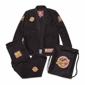 Puede incluir: Un conjunto de uniforme de artes marciales negro, que incluye una chaqueta, pantalones y una bolsa con cordón. El uniforme presenta parches bordados dorados y morados con el texto "Shoyoroll". La chaqueta y los pantalones están extendidos, y la bolsa está de pie.