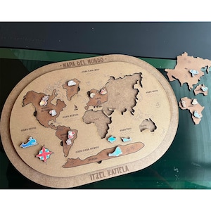 Op de afbeelding: Een houten wereldkaartpuzzel met continenten en oceanen in het Spaans. Inclusief verwijderbare continentstukken en decoratieve elementen. De tekst "MAPA DEL MUNDO" en "ITZEL KATIELA" zijn zichtbaar.