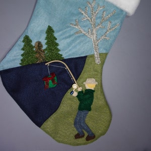 Op de afbeelding: Een blauwe en groene kerstsok met een witte boord. De sok heeft een geborduurde sc&egrave;ne van een man die aan het vissen is in een meer met een boom en een kale boom op de achtergrond. De man draagt een groen jasje en een blauwe broek. Een rood en groen cadeau hangt aan de hengel.
