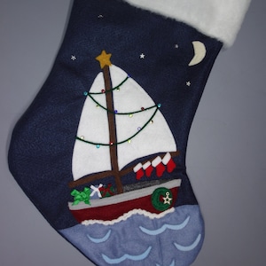 May include: A blue Christmas stocking with a white sailboat design. The sailboat has a brown mast, a white sail, and a red hull. The sailboat is decorated with a green Christmas tree, red and white stockings, and a green wreath. The stocking is decorated with white stars and a crescent moon.
