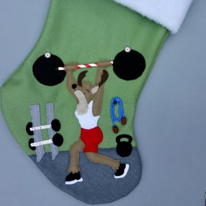 May include: Green Christmas stocking with a white cuff. The stocking features a brown reindeer lifting a barbell with red and white striped weights. The reindeer is wearing a white tank top and red shorts. There are other gym equipment items on the stocking, including dumbbells, a jump rope, and a kettlebell.