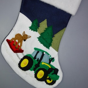 May include: A blue and white felt Christmas stocking with a green tractor, a brown deer, and green trees on a white background.