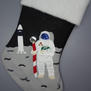 May include: A black and white Christmas stocking with a moon landing theme. An astronaut in a white spacesuit holds a candy cane and stands next to a rocket ship on the moon's surface.