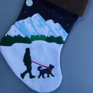 May include: A blue and white Christmas stocking with a winter scene of a person walking a dog in front of a mountain range under a full moon. The stocking has a brown cuff.