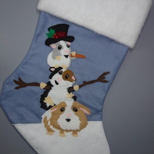 May include: A blue Christmas stocking with a white cuff and toe, decorated with a snowman made of guinea pigs. The snowman wears a black top hat and holds a carrot. Brown stick arms extend from the middle guinea pig.