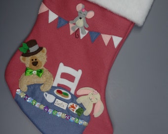 May include: Pink felt Christmas stocking with a white faux fur cuff. The stocking features a scene of a bear, bunny, and mouse having tea at a table with a chair. The table is decorated with a green bow, a plate of food, and a teacup. The stocking is decorated with a string of iridescent sequins and the name "Santa" is written on the table.