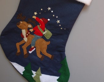 Op de afbeelding: Een blauwe kerstsok met een witte boord. De sok heeft een vilt applicatie van een rendier en een persoon die op het rendier rijdt. De scène speelt zich af tegen een donkerblauwe achtergrond met gouden glittersterren.