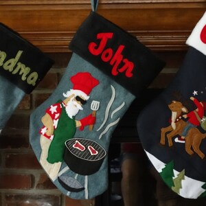 May include: Three Christmas stockings hang on a mantel. Each stocking is denim blue with a black cuff and a name embroidered in red or green. One stocking has a snowman, another has a grilling Santa, and the last has a child riding a reindeer.