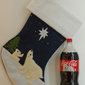 Op de afbeelding: Een blauwe kerstsok van vilt met een witte boord en een witte applikatie van een ijsbeer. De sok heeft een witte ster en een groene dennenboom-applikatie. De sok ligt naast een fles Coca-Cola.
