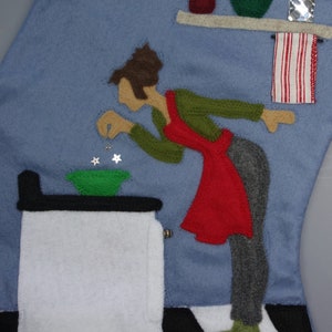 May include: A felt Christmas stocking depicting a kitchen scene. A figure in a red apron and green shirt is adding stars to a green bowl. A white stove, a shelf with utensils, and a red and white striped towel are also featured.