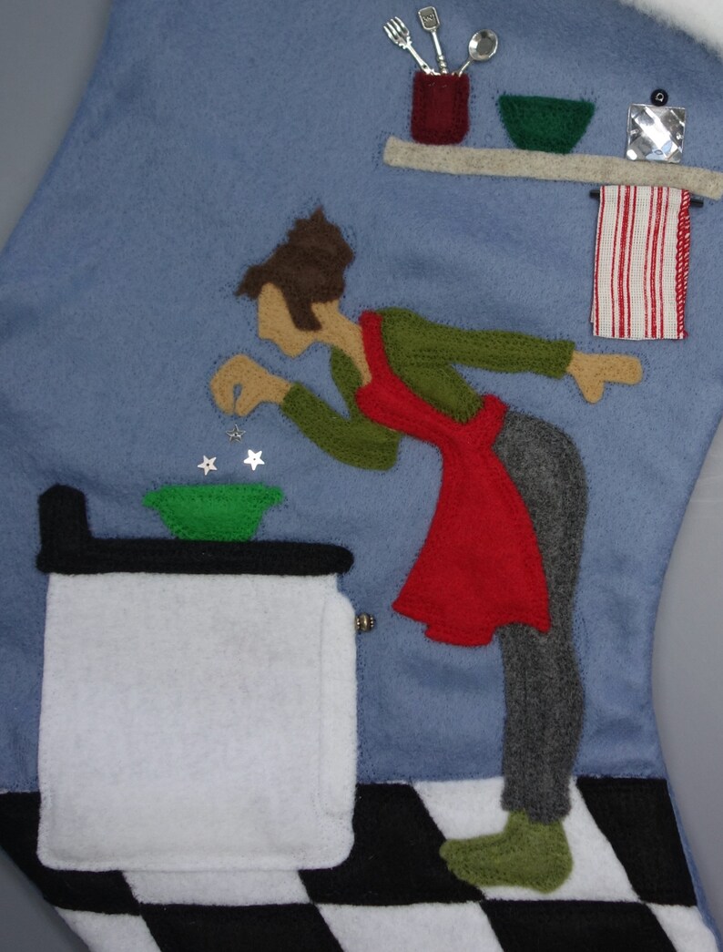 May include: A felt Christmas stocking depicting a kitchen scene. A figure in a red apron and green shirt is adding stars to a green bowl. A white stove, a shelf with utensils, and a red and white striped towel are also featured.