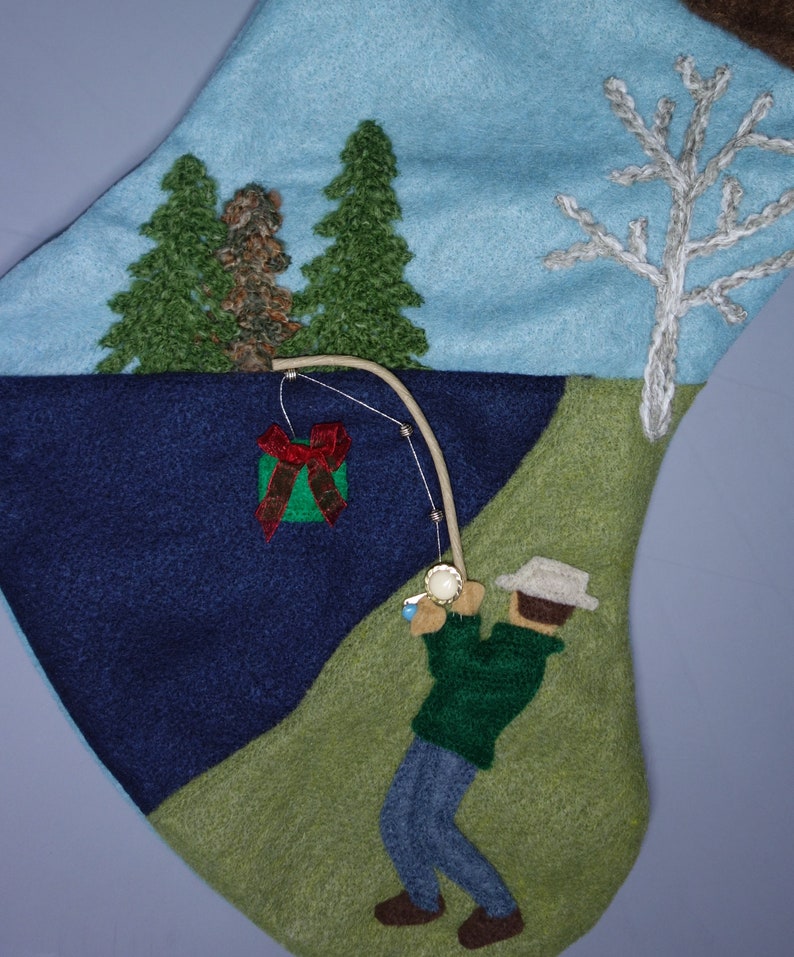 Op de afbeelding: Een blauwe, groene en bruine wollen kous met een wintersc&egrave;ne. De kous toont een visser met een hengel, een cadeaudoos en twee dennenbomen.
