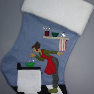 May include: A blue Christmas stocking with a white cuff. The stocking features an applique of a person in a red apron, green shirt, and grey trousers, cooking in a kitchen. The kitchen has a black and white chequered floor.