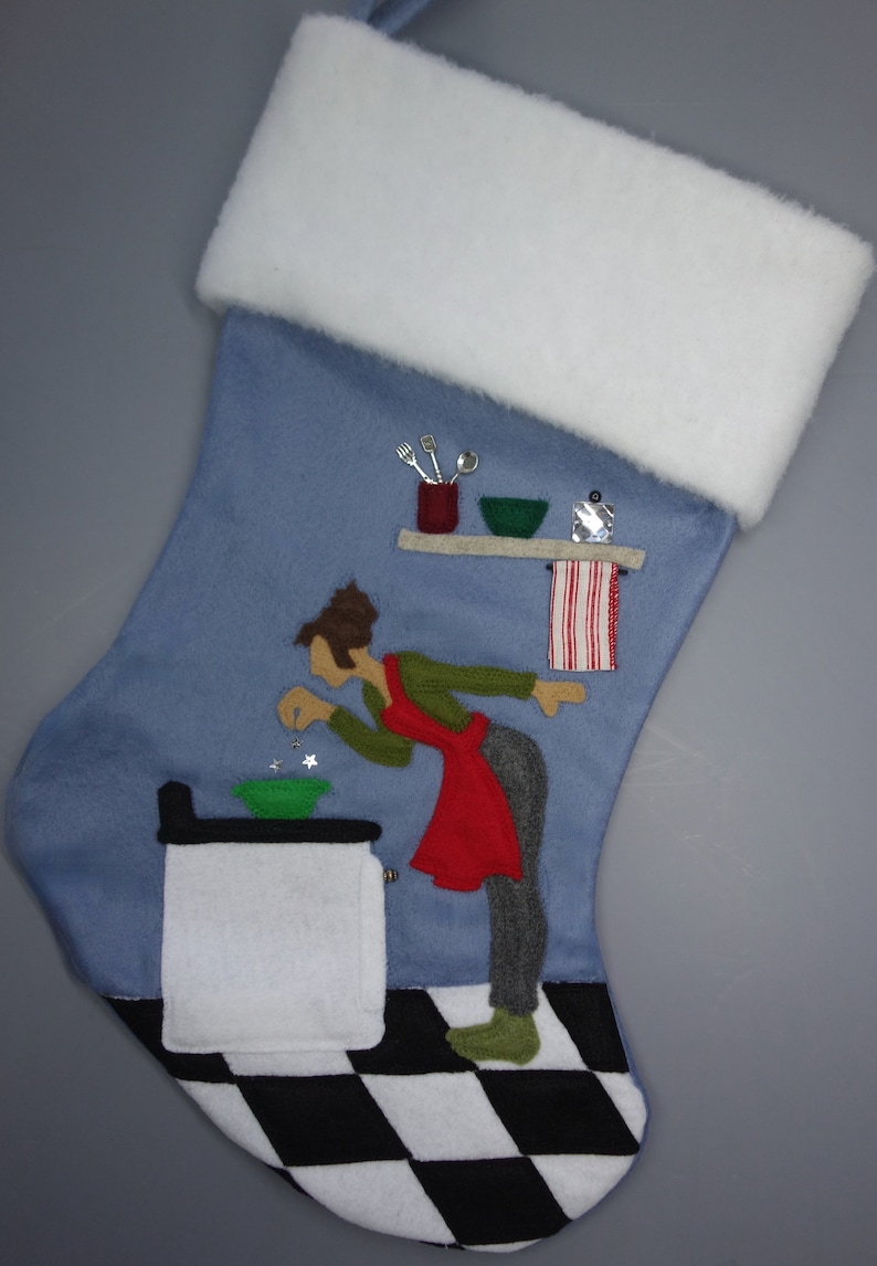 May include: A blue Christmas stocking with a white cuff. The stocking features an applique of a person in a red apron, green shirt, and grey trousers, cooking in a kitchen. The kitchen has a black and white chequered floor.