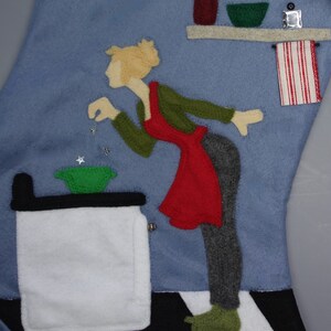 May include: A blue felt Christmas stocking with a kitchen scene. A woman in a red apron and green top is reaching towards a green bowl. The scene includes a stove, shelf with utensils, and a red and white striped towel.