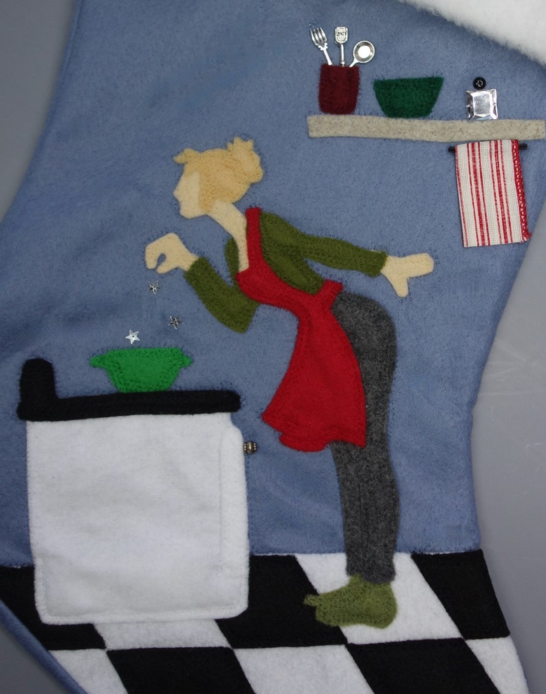 May include: A blue felt Christmas stocking with a kitchen scene. A woman in a red apron and green top is reaching towards a green bowl. The scene includes a stove, shelf with utensils, and a red and white striped towel.