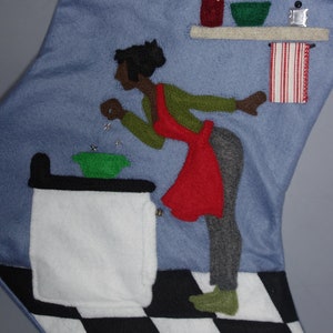 May include: Blue felt Christmas stocking with a kitchen scene. A woman in a red apron and green sweater is depicted near a stove. The stocking has a black and white checkered floor design.