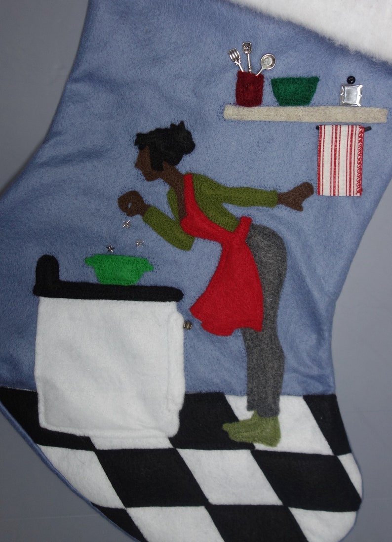 May include: Blue felt Christmas stocking with a kitchen scene. A woman in a red apron and green sweater is depicted near a stove. The stocking has a black and white checkered floor design.
