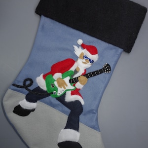 May include: Blue Christmas stocking featuring a felt appliqué of Santa Claus playing an electric guitar. Santa is wearing sunglasses and a red suit.