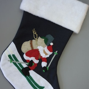 May include: A blue and white Christmas stocking featuring a felt appliqué of Santa Claus skiing down a snowy slope. Santa is wearing a red suit, white beard, and black boots. He is holding ski poles and has a bag of gifts on his back.