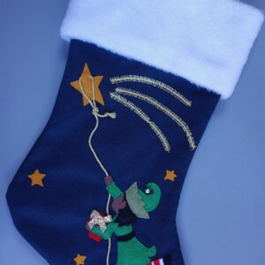 May include: A blue Christmas stocking with a white cuff. The stocking features a green elf holding a gift and climbing a rope attached to a gold star. The background is a dark blue with gold stars.