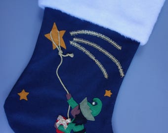 Op de afbeelding: Een blauwe kerstsok met een witte boord. De sok heeft een groene elf die een cadeau vasthoudt en klimt aan een touw dat vastzit aan een gouden ster. De achtergrond is donkerblauw met gouden sterren.