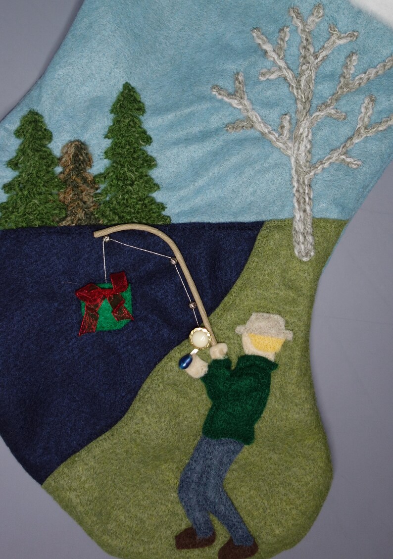 Op de afbeelding: Een wollen kous met een blauwe en groene landschapssc&egrave;ne. Een man met een hoed vist met een hengel en een molen. Een groene geschenkdoos zit aan de lijn. De kous is versierd met een witte boom en twee groene bomen.