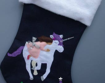 Op de afbeelding: Een zwarte kerstsok van vilt met een witte boord. De sok heeft een wit eenhoorn met een paarse manen en hoorn, een meisje dat op de eenhoorn rijdt, en een kasteel met roze en paarse accenten.