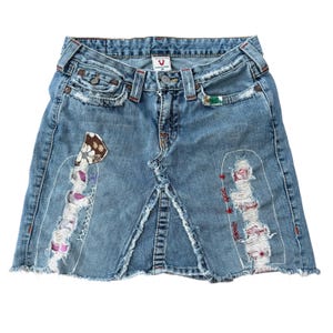 Könnte beinhalten: Ein hellblauer Jeans-Minirock mit ausgefranstem Saum, abgenutzten Partien mit sichtbaren weißen Nähten und dekorativen Stoffflicken mit Blumen- und Mustermotiven. Das Kleidungsstück hat einen Knopfverschluss und ein True Religion Markenetikett.
