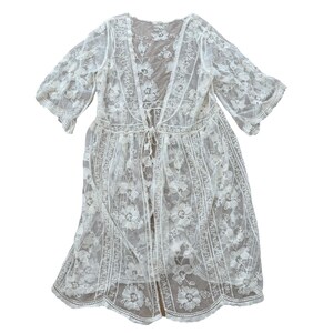 May include: A sheer white lace duster featuring an intricate floral pattern, three-quarter length sleeves, and a delicate tie-front closure. The garment has a scalloped hemline and vertical panels of embroidery that extend from the neckline to the bottom.